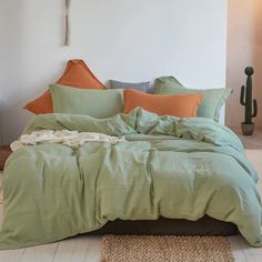 a bed with green sheets and orange pillows in a white room next to a cactus