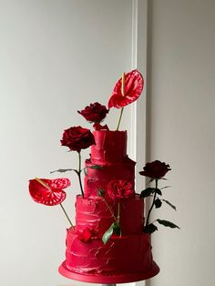 a three tiered cake with red icing and flowers on top, sitting in front of a white wall