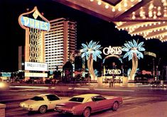 two cars are parked in front of the casino at night time with lights on and palm trees lit up