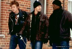 three men walking down the street in front of a brick building, one wearing a beanie