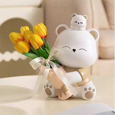 a white teddy bear holding yellow tulips on top of a table next to a vase