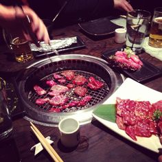 some food is being cooked on a grill with chopsticks next to it and people sitting at a table in the background