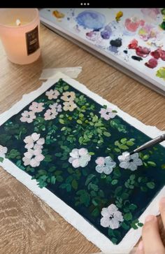 a person is painting flowers on a piece of paper with a candle in the background