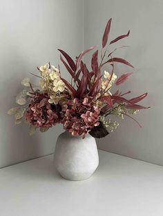 a white vase filled with lots of flowers on top of a table next to a wall