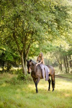 a woman riding on the back of a brown horse through a lush green forest filled with trees