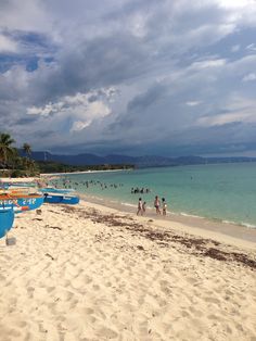 many people are on the beach with their surfboards and boats in the water under a cloudy sky