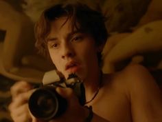 a shirtless young man holding a camera in his hand and looking at the camera