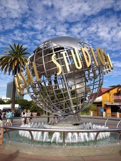 people are walking around the fountain in front of a globe with words that spell out my studio on it