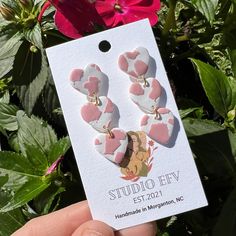 someone is holding up some pink and white heart shaped earrings in front of red flowers