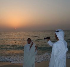 two people standing on the beach at sunset with one holding a camera to take pictures