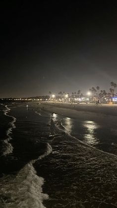 the beach at night with lights in the distance and people walking on the shore line