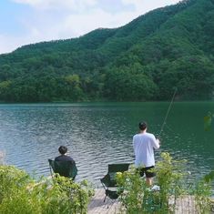 two people sitting on chairs fishing from a dock near a lake with mountains in the background