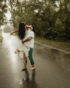 a man and woman dancing in the middle of the road on a rainy day with trees behind them