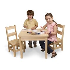 two children sitting at a wooden table and chair with their hands on the paper that they are writing