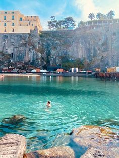 a person swimming in the water near some rocks and buildings on top of a hill