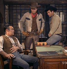 three men sitting around a table in an old time western town setting with one man pointing at another man