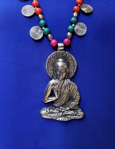 Stay calm and in the Zen wearing this large hand crafted Buddha pendant in oxidized silver, strung on a necklace of colorful beads and discs. Buddha poses have different meanings. There are over 100 hand gestures, or postures, called mudras. Here the Buddha is in the abhaya gesture, with right hand raised, fingers upwards, palm facing out, while the left hand is next to the body. This mudra is the gesture of fearlessness. Wear it and be fearless On a long necklace of colored beads, silver color Hand-strung Long Necklace For Meditation, Buddhism Necklace, Yoga Jewelry Buddha Groove, Hand-strung Pendant Jewelry For Meditation, Buddha Gifts, Buddah Necklaces, Zen Jewelry, Resort Jewelry, Festival Necklace