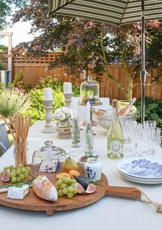 an outdoor table set with plates and wine glasses, grapes, cheese, bread, and other food items