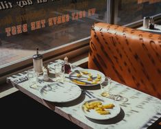 two plates of food on a table in front of a window with an orange chair