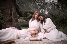 three women in white dresses are laying on a blanket under a tree and posing for the camera