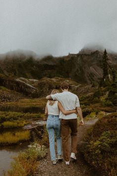 a man and woman walking down a path in the mountains with their arms around each other