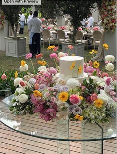 a table topped with a white cake surrounded by lots of flowers and people in the background