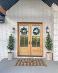 two wreaths are on the front door of a white brick house with potted plants