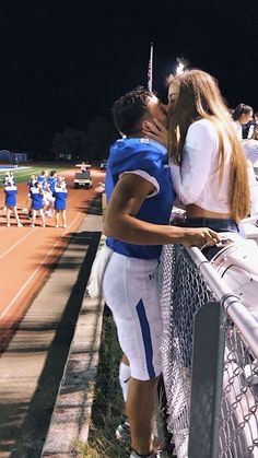 a man and woman kissing on the side of a fence at a football game with other people in the background