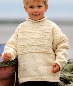 a little boy standing next to a wooden barrel on the beach wearing a white sweater and black pants