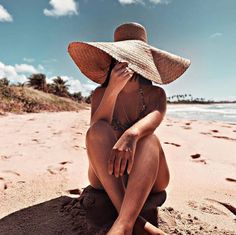 a woman in a hat sitting on the beach with her legs crossed and hands behind her head