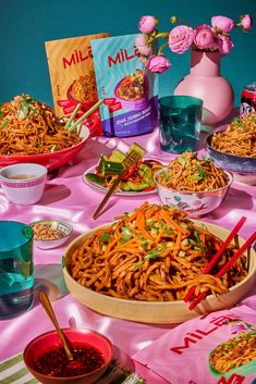 a table topped with lots of food next to bowls of noodles and sauces on top of a pink table cloth