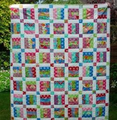 a colorful quilt hanging from the side of a fence in front of some trees and bushes