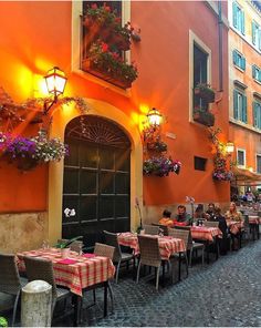 people are sitting at tables in an alleyway with flowers hanging from the building's windows