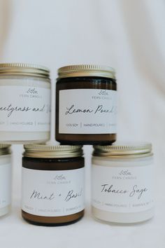 four jars of candles sitting next to each other on a white tablecloth covered surface