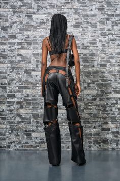 a woman in black leather pants and boots standing against a brick wall with her back to the camera