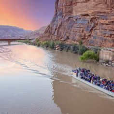 a long boat filled with people on the side of a river next to a bridge