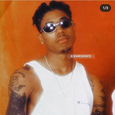 a man with tattoos and sunglasses standing in front of an orange wall wearing a white tank top