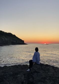 a man sitting on the edge of a cliff looking out at the ocean and sunset