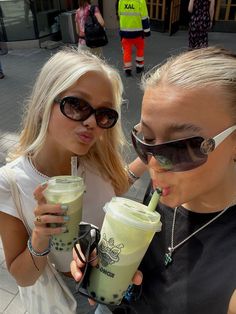 two young women are drinking iced coffee outside on the sidewalk while wearing sunglasses and holding drinks in their hands