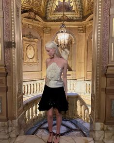 a woman in a white top and black skirt standing on a marble floor next to a chandelier