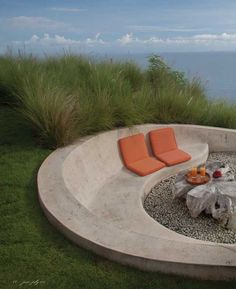 an outdoor fire pit surrounded by grass and rocks with two orange cushions on the seat