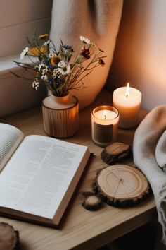 an open book on a table next to flowers and candles with a blanket over it