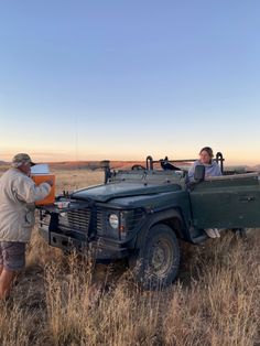 two men standing next to a truck in the middle of a dry grass covered field