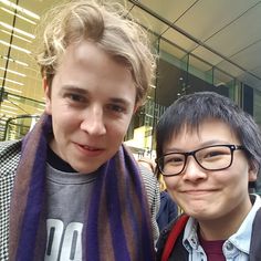 two young men standing next to each other in front of a glass building and one is wearing a scarf