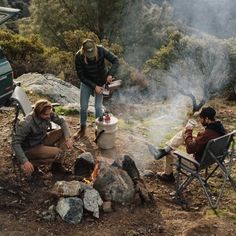 three people sitting around a campfire with food on the grill and one person standing next to it