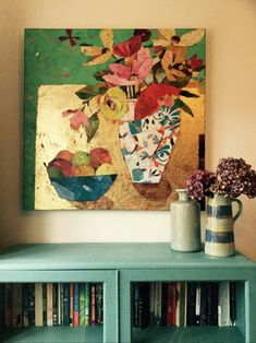 a painting is hanging on the wall above a book shelf with vases and flowers