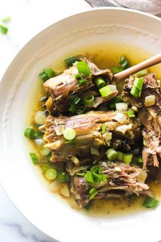 a white bowl filled with meat and vegetables on top of a marble table next to a wooden spoon