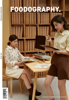 two young women sitting at a table in front of bookshelves, one holding a clipboard and the other reading