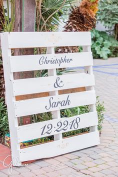 a wooden sign sitting on the side of a road next to a palm tree and potted plant