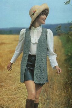 a woman walking in a field wearing a hat and vest with crochet on it